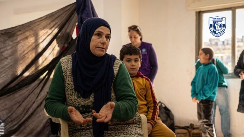 A middle-aged woman in a hijab and green sweater sitting in a displacement shelter, gesturing with her hands while speaking about the situation of women and girls in Southern Lebanon.